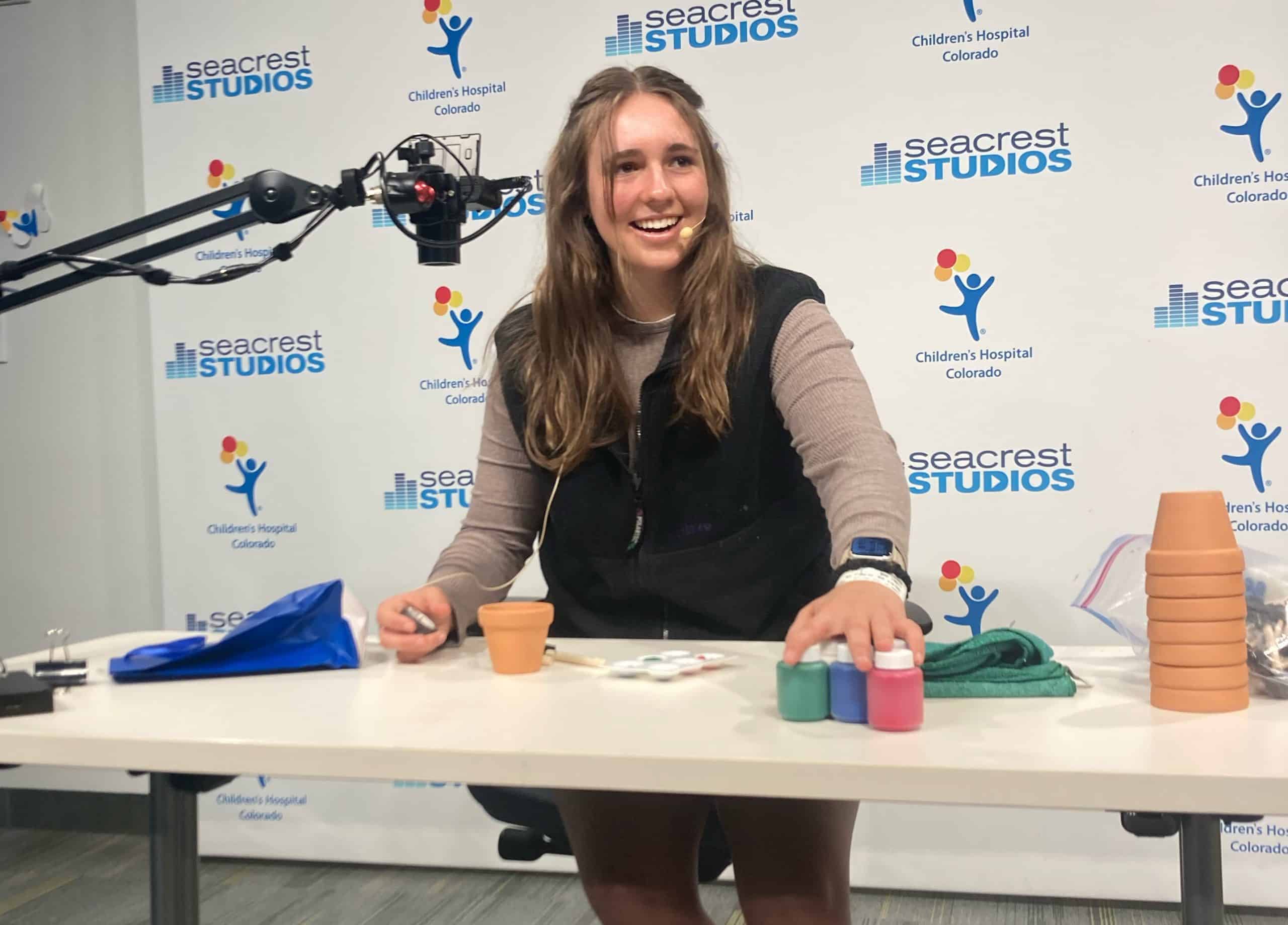 High school senior Elizabeth Marr is sitting at a desk in a studio at Children's Hospital. She is looking at the camera and has craft supplies on the table in front of her. There is a mic on a long stand in next to her face.
