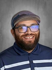 Smiling man with a beard and glasses, wearing a striped shirt and a cap, posed against a neutral background.