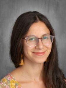 A smiling woman with long dark hair and glasses, wearing a floral top and tassel earrings, against a gray background.