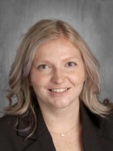Portrait of a woman with wavy, light hair, smiling, wearing a black blazer, against a neutral backdrop.