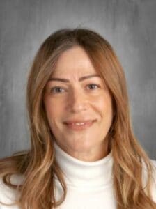 A woman with long, wavy hair wearing a white turtleneck smiles in front of a gray backdrop.