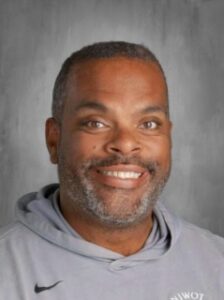 A smiling man with short gray hair and a beard, wearing a gray hoodie with a logo, against a light background.