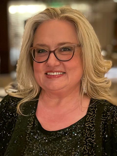 A smiling woman with long, wavy hair, wearing glasses and a sparkling black top, stands in a softly lit setting.