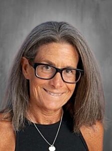 A smiling woman with long gray hair and glasses, wearing a black top and a silver necklace, against a gray background.