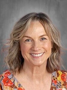 Smiling woman with shoulder-length blonde hair, wearing a colorful floral shirt, against a gray background.