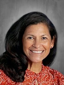 Smiling woman with long dark hair wearing a red patterned blouse, set against a gray background.