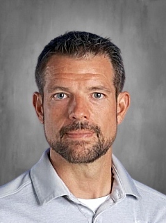 A man with short, dark hair and a beard, wearing a gray polo shirt, against a neutral background.