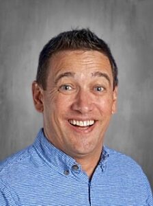 Smiling man with short brown hair wearing a blue and white striped shirt, set against a gray background.