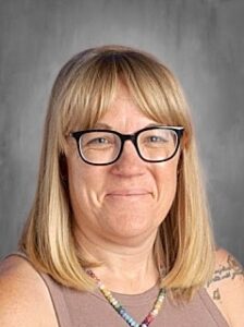 A smiling woman with shoulder-length blonde hair and glasses, wearing a beige top and colorful necklace.