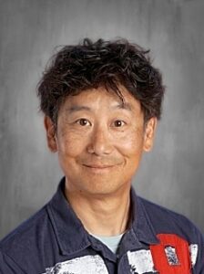 A smiling man with dark, wavy hair wearing a navy blue shirt with a colorful design against a gray background.