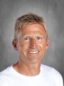 Smiling man with short blonde hair, wearing a white shirt, set against a light gray background.