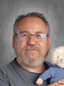 A man with glasses and a beard smiles while holding a plush toy resembling a character with white hair.