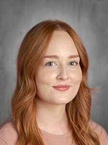 A woman with long red hair smiles at the camera, wearing a light pink top against a gray background.