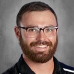 Smiling man with glasses and a beard, wearing a black shirt with white stripes, against a gray background.