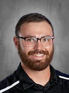 Smiling man with glasses and a beard, wearing a black shirt with white stripes, against a gray background.
