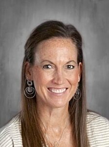 A smiling woman with long hair and earrings, wearing a light sweater, against a neutral background.