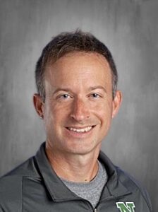 Smiling man with short brown hair wearing a gray athletic jacket, set against a plain gray background.