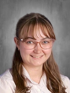 A young woman with glasses and long, wavy hair smiles at the camera, wearing a white blouse against a gray background.