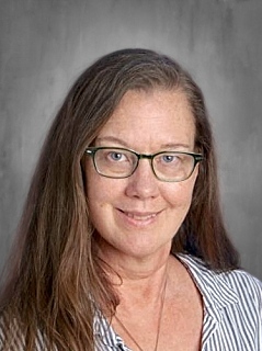 A woman with long brown hair and glasses smiles at the camera, wearing a striped shirt against a gray background.