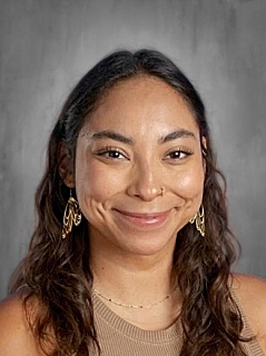 A smiling woman with curly hair, wearing gold earrings and a tan top, against a gray background.