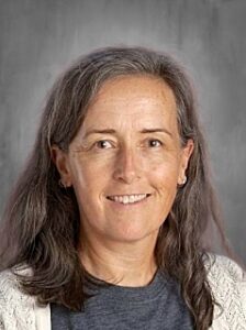 Portrait of a woman with long, gray hair, smiling, wearing a gray shirt and a light cardigan against a gray background.