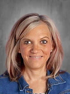 A woman with light brown hair, wearing a denim jacket, smiles against a gray background.