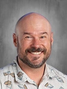 Smiling man with a bald head and a beard, wearing a patterned shirt with fish designs, against a gray background.