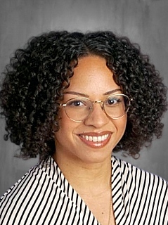 A smiling woman with curly hair and glasses, wearing a striped top, poses against a gray background.