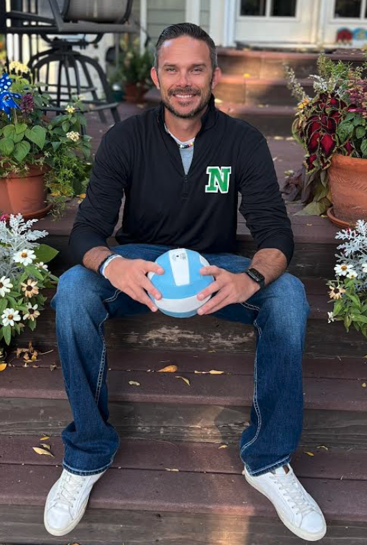 Coach sits on wooden steps surrounded by plants, holding a volleyball, wearing a black shirt with a green letter 'N'.