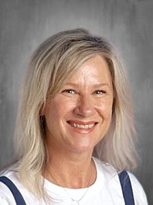 Smiling woman with shoulder-length blonde hair, wearing a white shirt and dark straps, against a gray background.