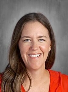 A smiling woman with long brown hair, wearing an orange top, against a neutral gray background.