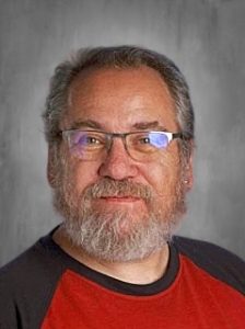 Man with glasses and a beard wearing a red and black shirt, smiling at the camera against a gray background.