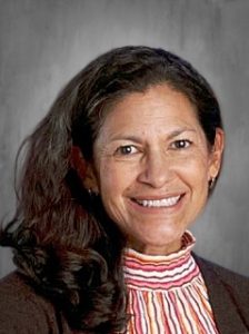 Smiling woman with long dark hair, wearing a striped turtleneck and brown jacket against a gray background.