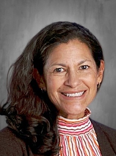 Smiling woman with long dark hair, wearing a striped turtleneck and brown jacket against a gray background.