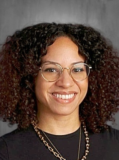 Persona sonriente con el pelo rizado, que lleva gafas y una camiseta negra con un collar de cadena dorada, sobre un fondo gris.