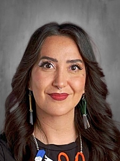 Mujer con el pelo largo y oscuro, con pendientes y un aro en la nariz, sonriendo sobre un fondo gris.