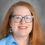 Smiling woman with glasses and long red hair, wearing a blue shirt and geometric earrings, against a gray background.