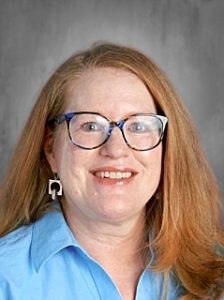 Smiling woman with glasses and long red hair, wearing a blue shirt and geometric earrings, against a gray background.
