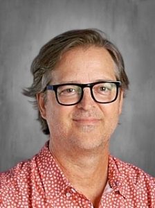 Man with glasses and short hair wearing a red patterned shirt, smiling in front of a gray background.
