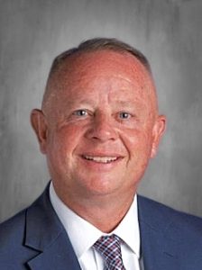 Man in a blue suit and tie smiling against a gray background.